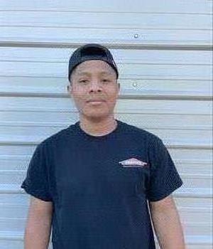 A Hispanic man with black shirt smiling in front of warehouse.